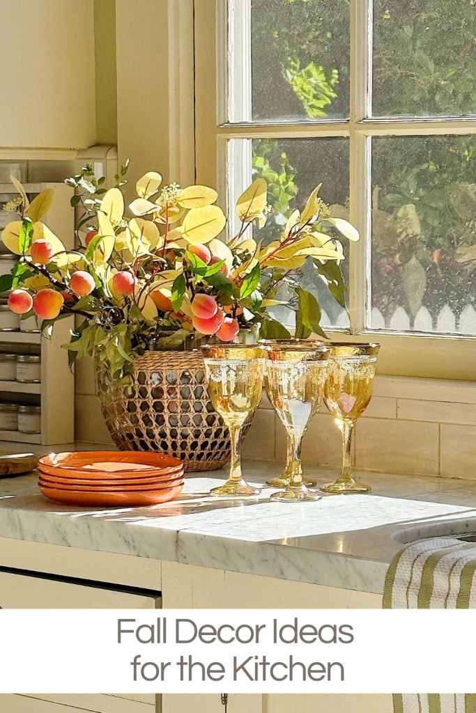 A kitchen countertop with fall-themed decor, including a basket of artificial leaves and fruit, three amber-tinted glasses, and orange plates. Sunlight streams through the window above.