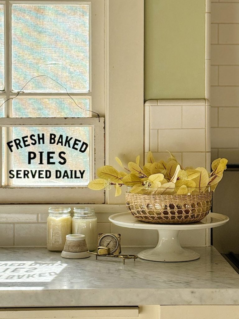 A kitchen counter features a basket with yellow leaves on a cake stand, two mason jars, a small clock, a candle, and a window sign reading "Fresh Baked Pies Served Daily.