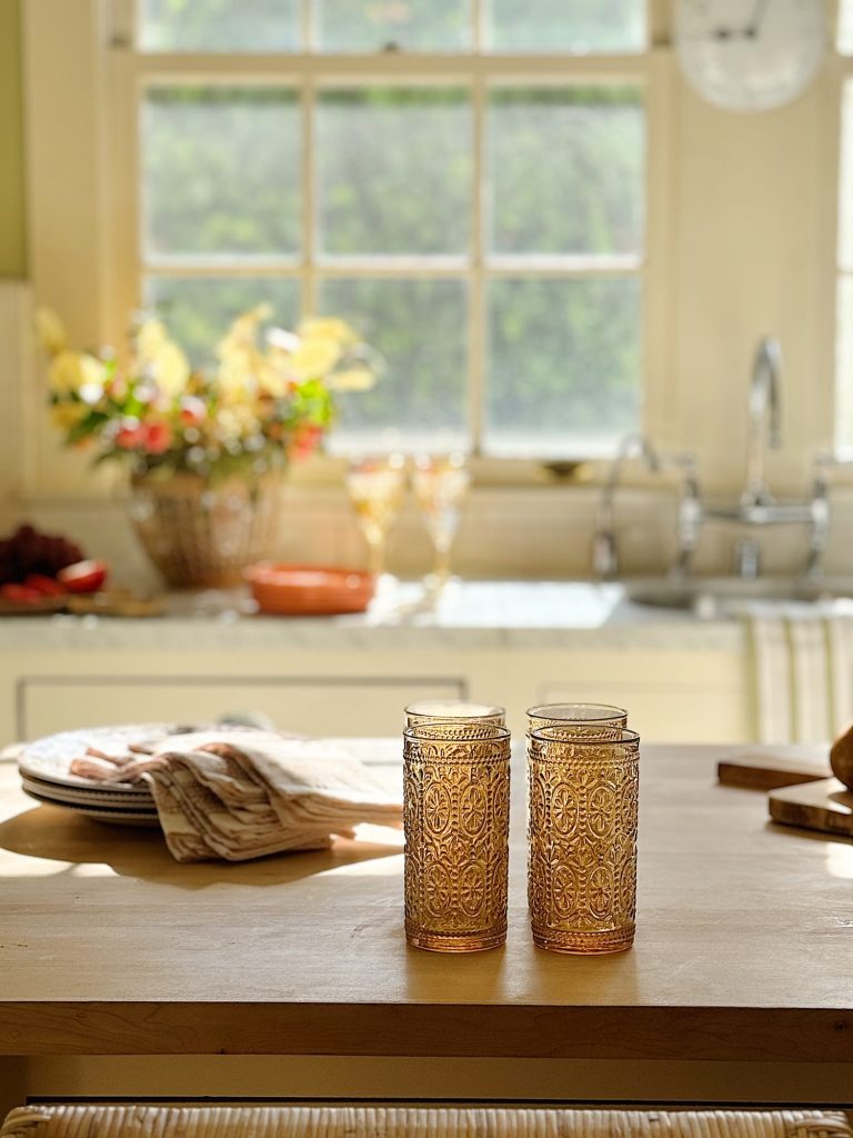 A kitchen with a wooden table featuring two ornate glasses, a stack of plates with cloth napkins, and a basket of flowers near a sunlit window.