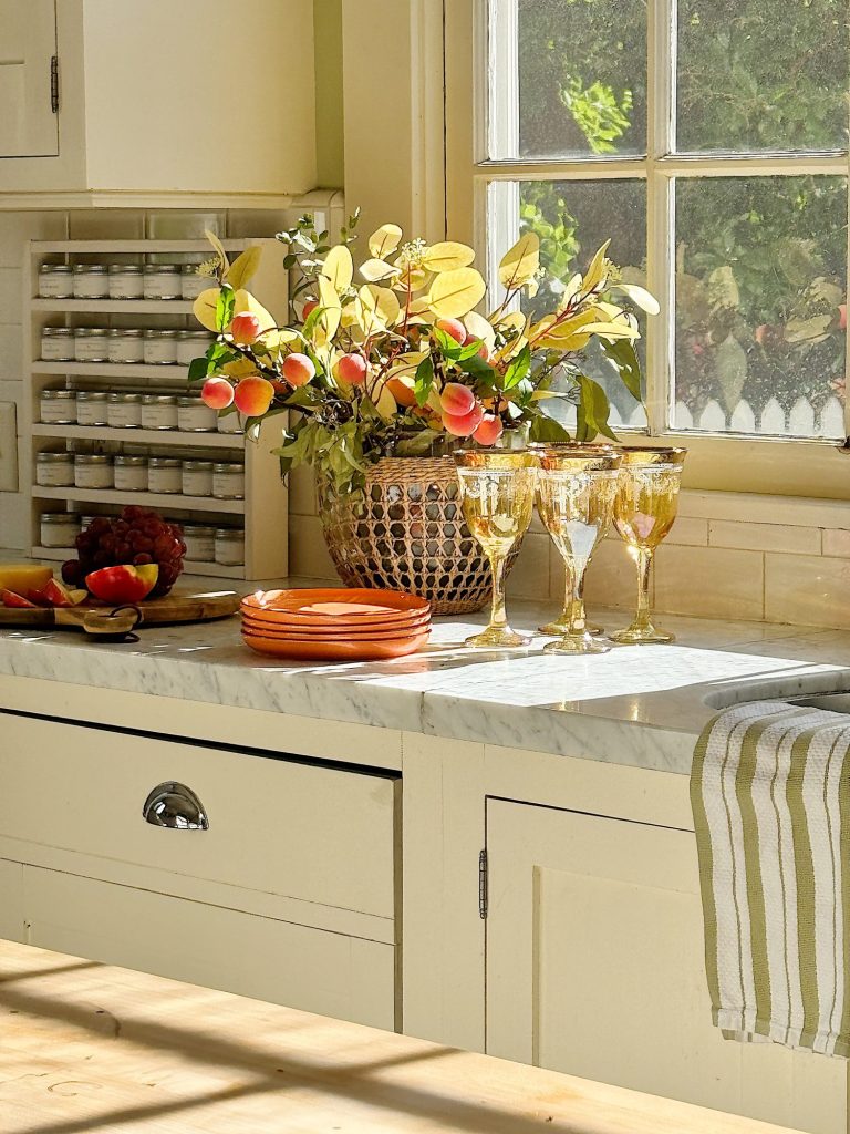 A sunlit kitchen counter with stacked orange plates, four gold-rimmed wine glasses, a fruit and foliage arrangement, and a spice rack next to a window with a striped towel hanging nearby.