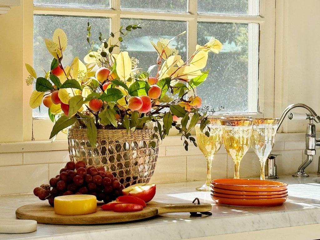 A kitchen counter with a fruit and leaf arrangement in a basket, four decorative goblets, a stack of orange plates, and a cheese board with cheese, apples, and grapes, all next to a window.