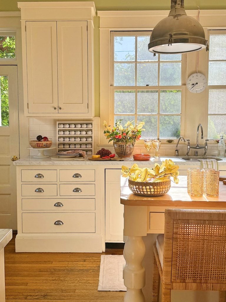 A bright kitchen features white cabinetry, a countertop with fruit bowls, a basket of eggs, wicker chairs, and sunflowers in a vase by large windows. A vintage-style light fixture hangs above the island.