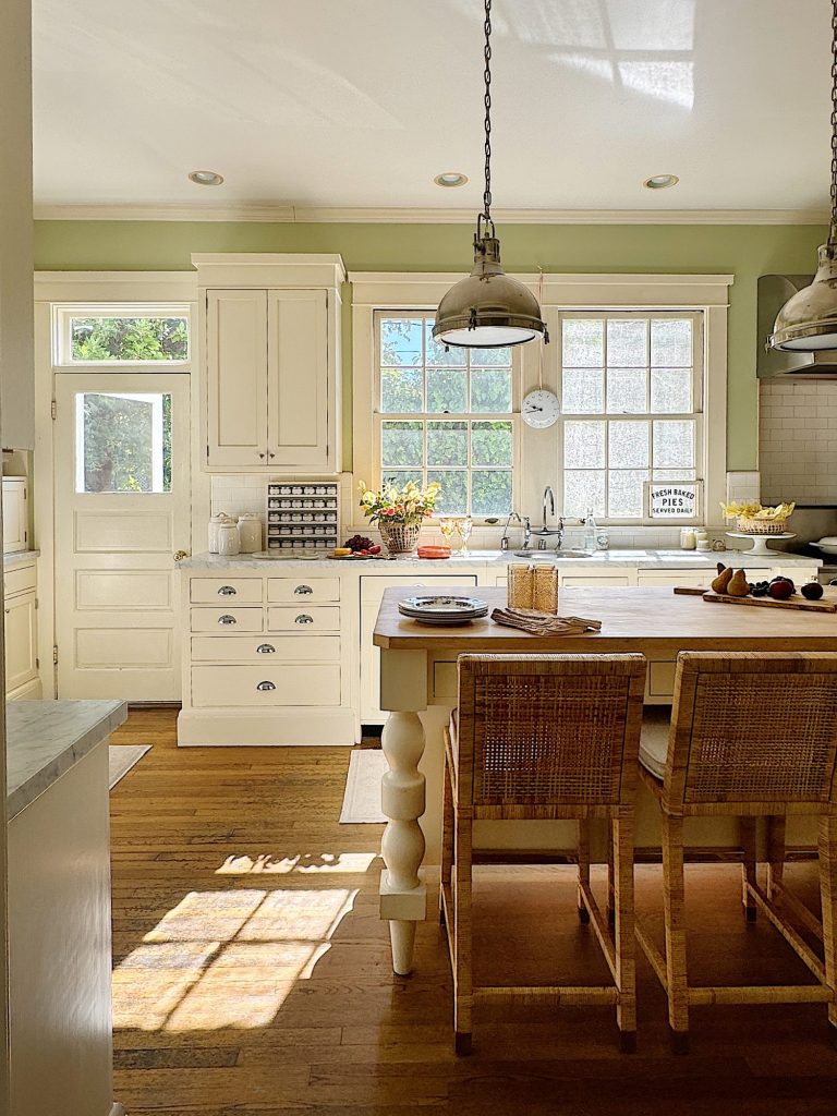 Sunny kitchen with white cabinets and island, wooden stools, pendant lights, and large windows. Plates and utensils are set on the island. There is a door and a window with a view of greenery outside.