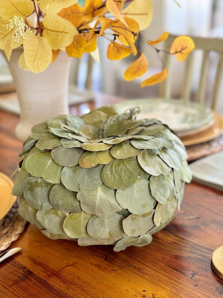 A decorative pumpkin made of leaves sits on a wooden table, surrounded by a vase of autumn-colored branches and place settings.