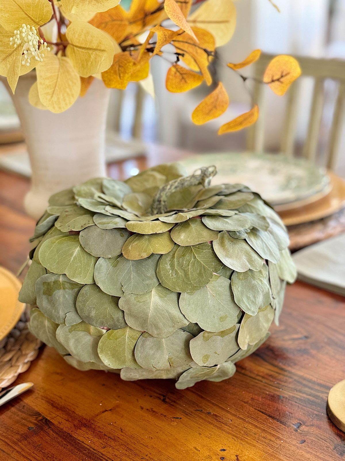A decorative pumpkin made of leaves sits on a wooden table, surrounded by a vase of autumn-colored branches and place settings.