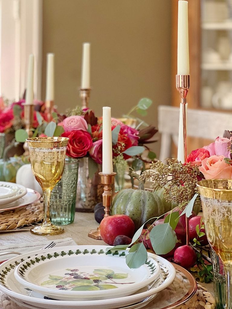 A dining table is set with floral arrangements, candles, and fruit in a fall-themed decor. Wine glasses and plates with flower designs are arranged neatly.