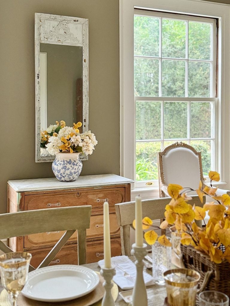 A dining room with a wooden table, decorated with white and yellow flowers, two lit candles, a vintage dresser with a mirror, and a large window showing greenery outside.