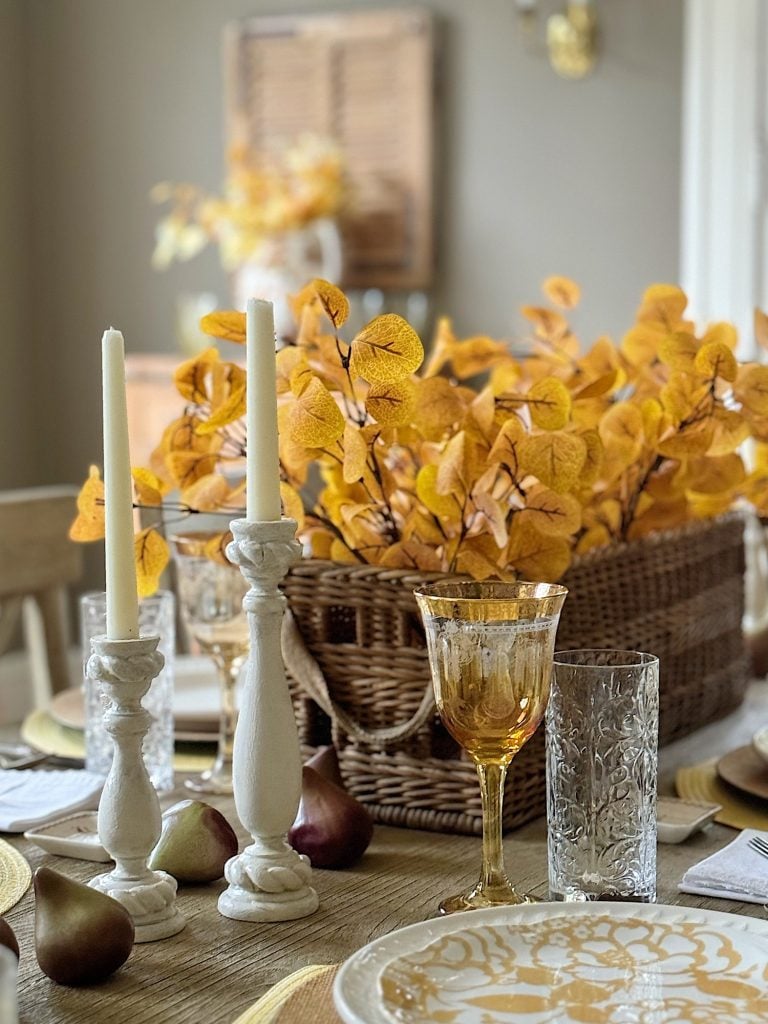 A dining table set with autumn-themed decor, including amber glassware, white candlesticks with lit candles, yellow foliage in a wicker basket, plates with gold patterns, and brown pears.