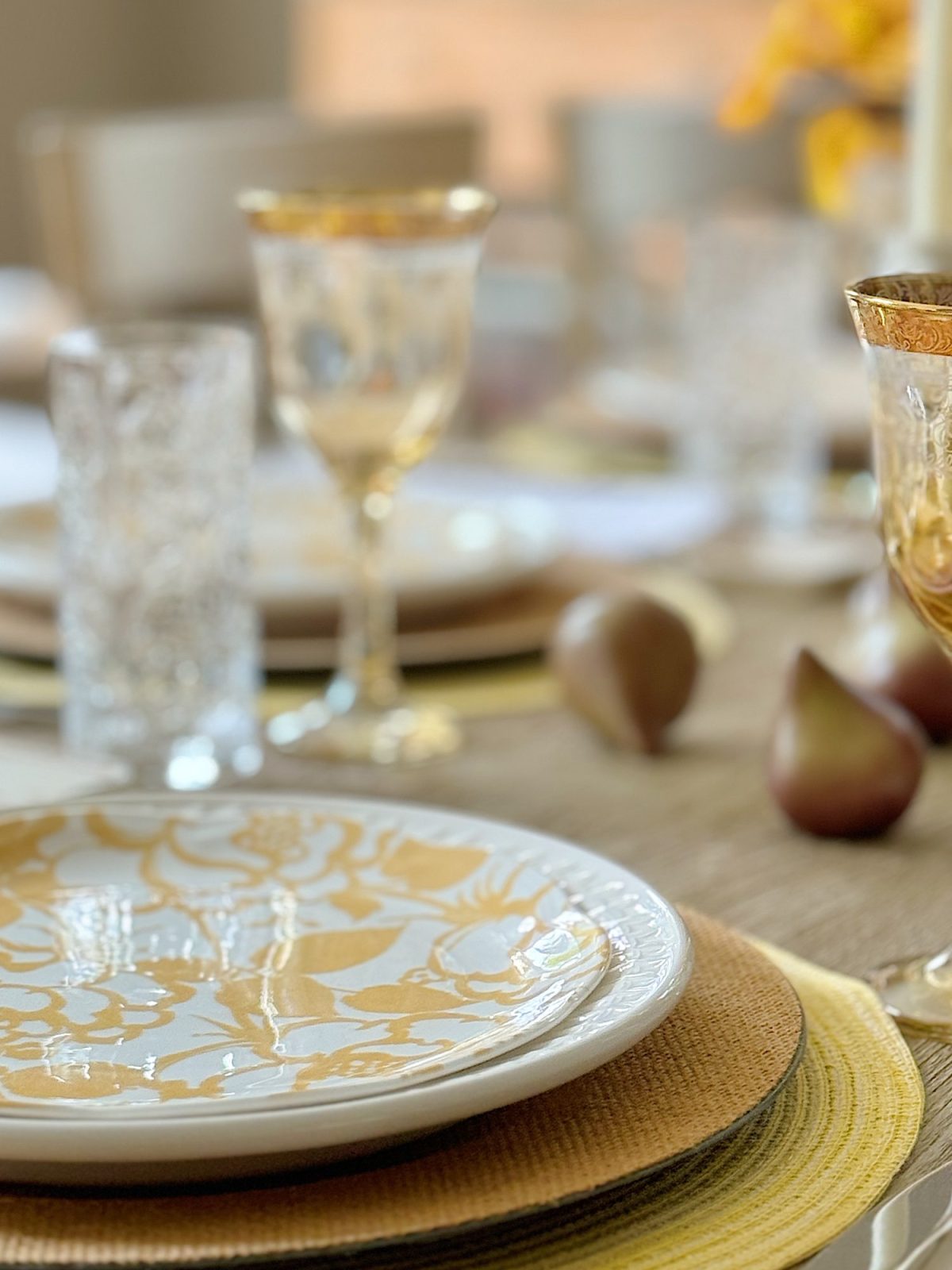 A close-up view of a dining table set with elegant gold-rimmed glassware and plates featuring ornate gold patterns. The table is adorned with a beige tablecloth and decorative items.