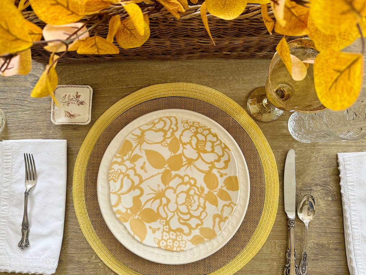 A place setting with a floral-patterned plate, yellow placemat, white napkin with a fork on the left, knife and spoon on the right, yellow leaves centerpiece, and a wine glass.