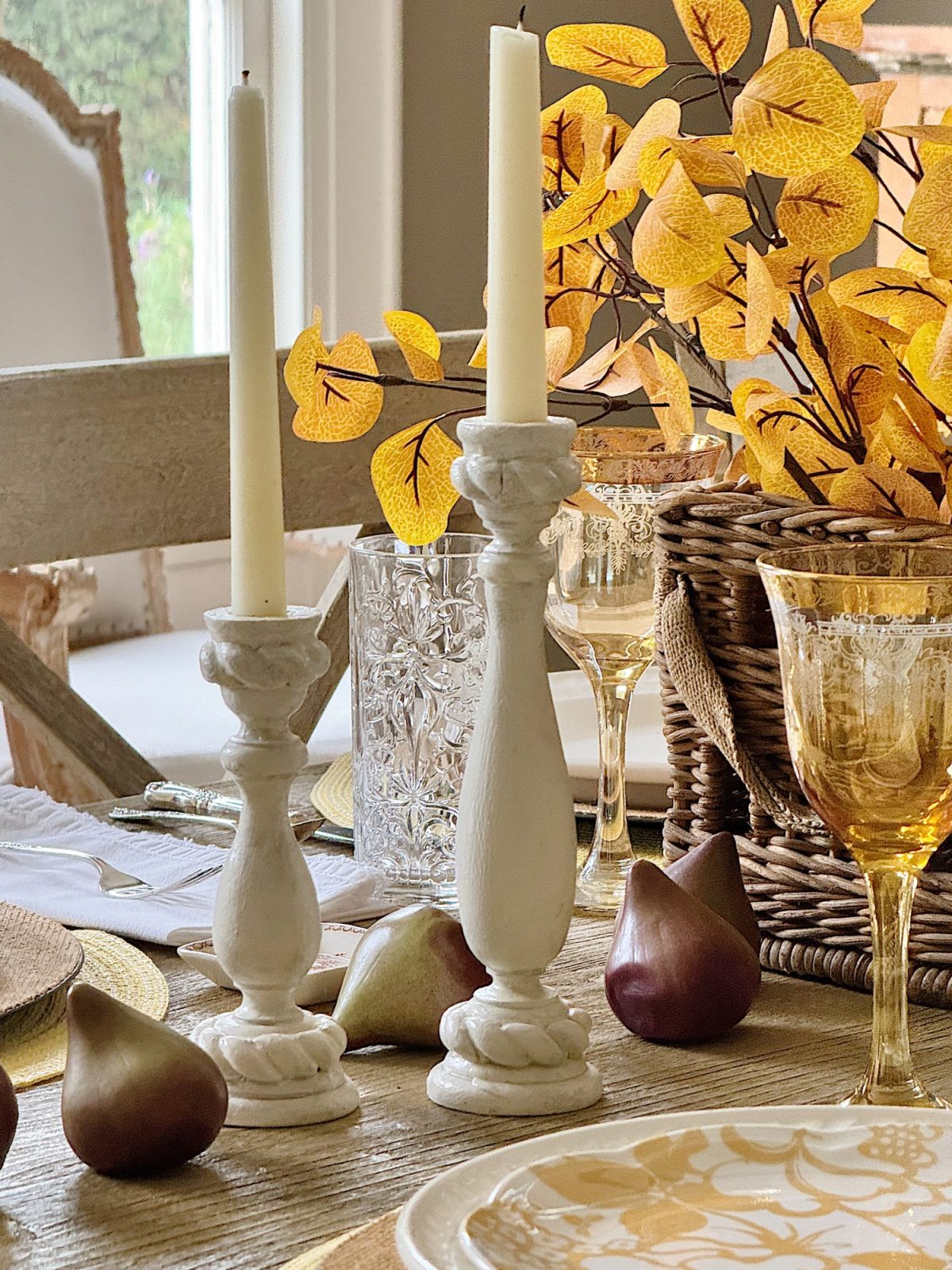 A dining table is decorated with two white candlesticks with lit candles, yellow-leafed branches in a woven basket, glasses, and pears.
