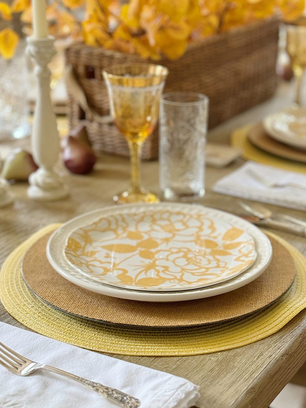A neatly arranged dining table with a yellow floral-patterned plate, glassware, and cutlery. Decorative yellow flowers and a wicker basket centerpiece are also present.