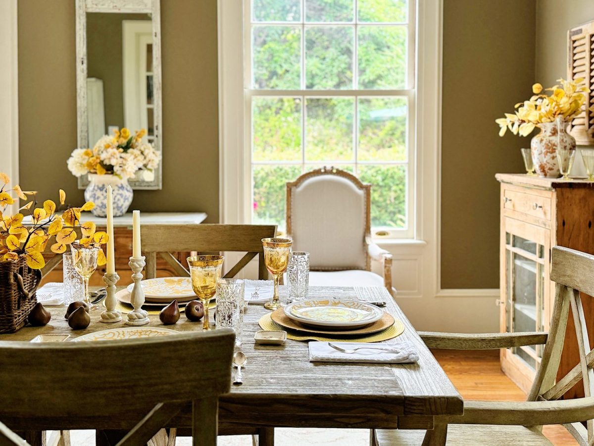 A dining area with a wooden table set for four. The setting includes plates, glasses, and a lit candle. The room features a large window, mirror, and floral arrangements. Warm, neutral tones are present.