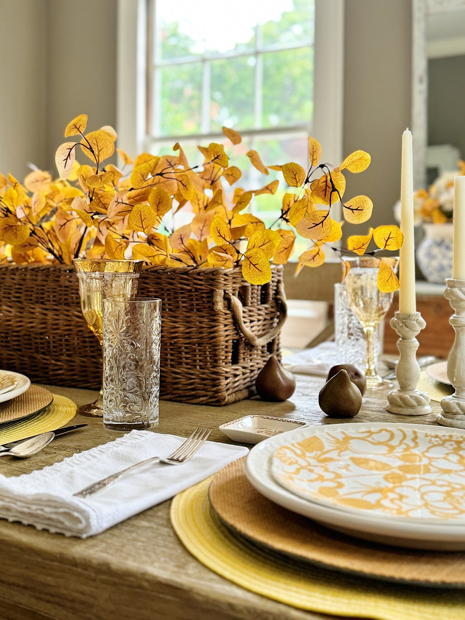 A dining table set with golden-themed plates, glassware, and napkins. There is a wicker basket with yellow foliage centerpiece, two candles, and decorative pears.