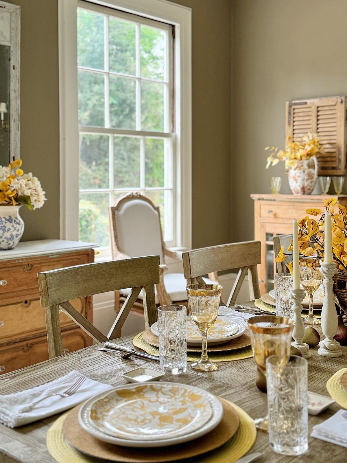 A dining room with a set table includes glassware, plates, and a centerpiece of flowers. The room has large windows, wooden furniture, and decor with a neutral color palette.