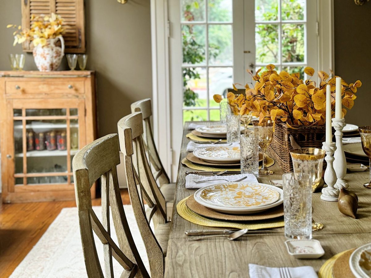 A dining room setup with a wooden table, decorated with yellow floral centerpiece, patterned plates, and crystal glasses. A wooden cabinet with glass doors stands in the background beside a window.