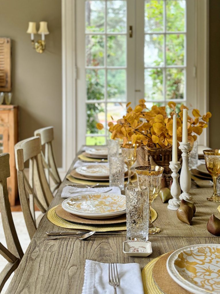 A dining table set for a meal with plates, glasses, cutlery, napkins, candle holders, and a centerpiece of golden leaves, against the backdrop of French doors leading to a garden.