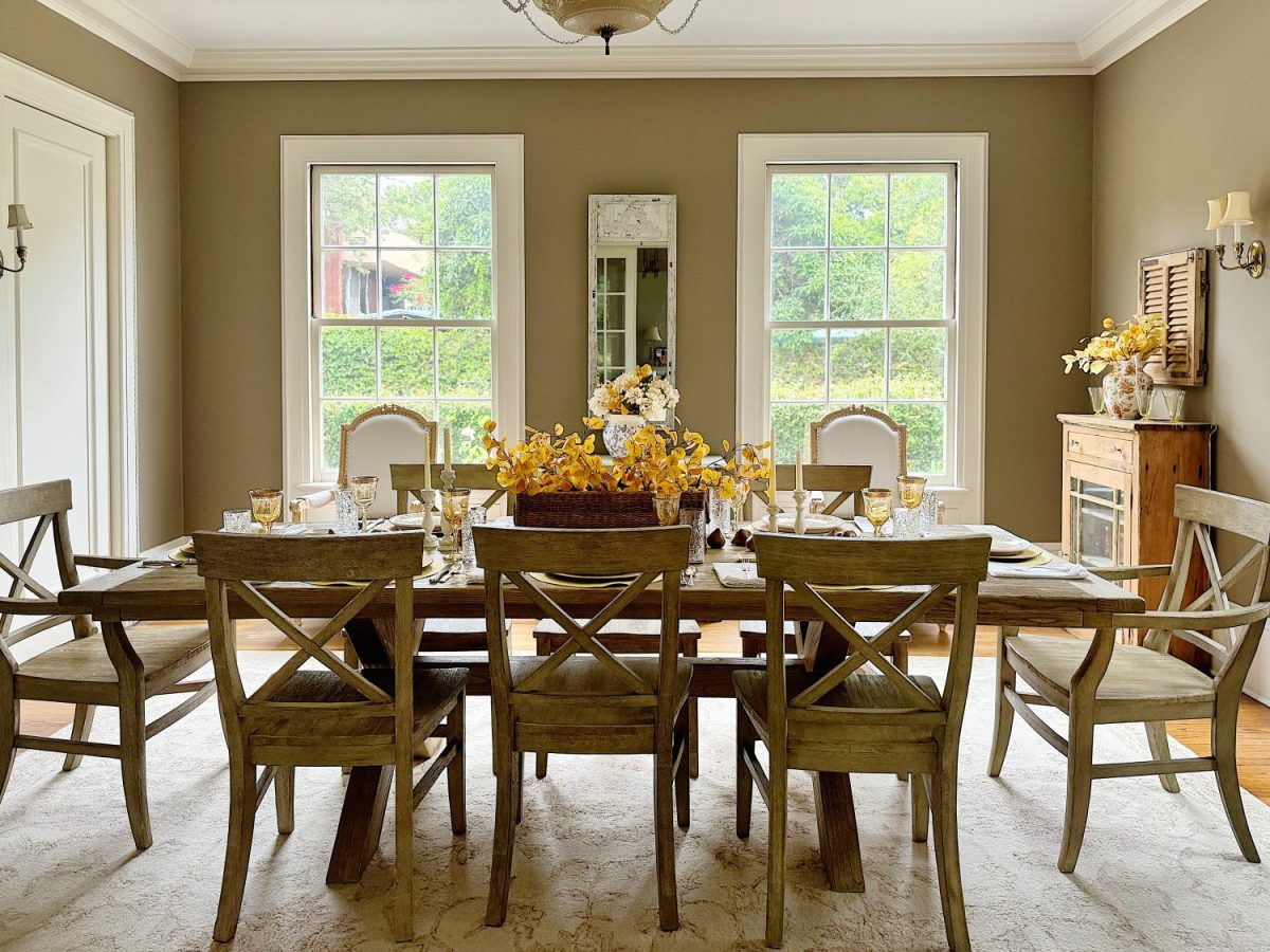 A dining room with a wooden table set for eight, featuring yellow floral centerpieces. Two large windows provide natural light, and the walls are painted light brown. Light wooden chairs surround the table.