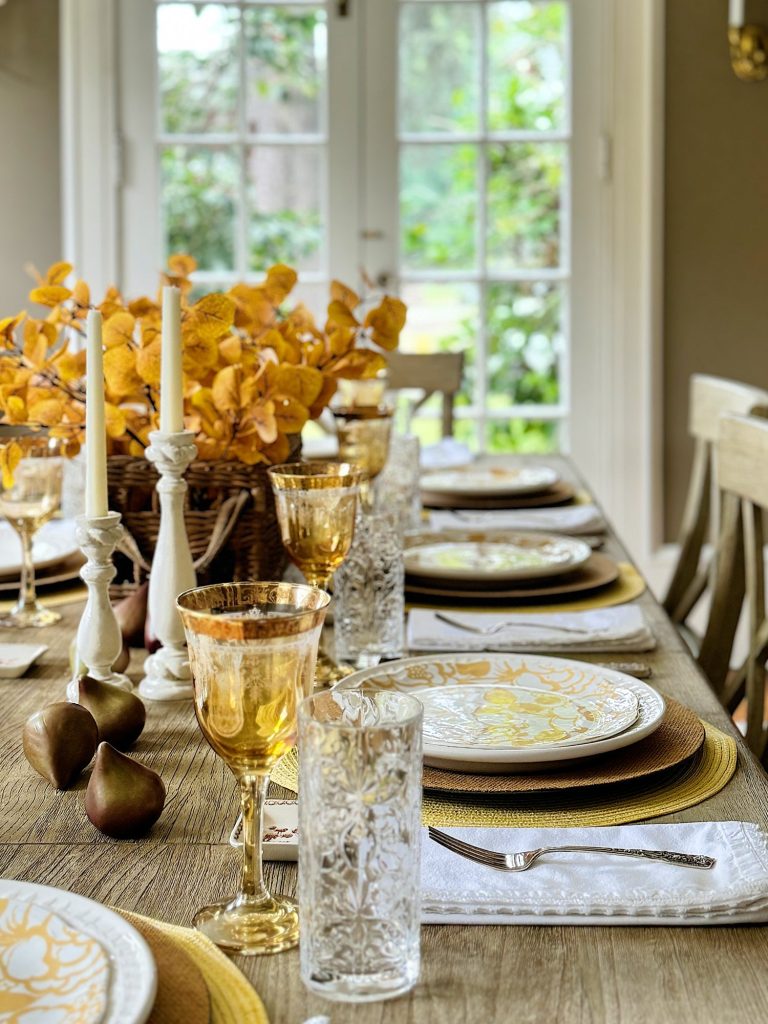 A dining table set for a meal with autumn-themed decor, featuring yellow floral plates, gold-rimmed glasses, white candles, and a centerpiece of yellow leaves and pears. The background shows glass doors.