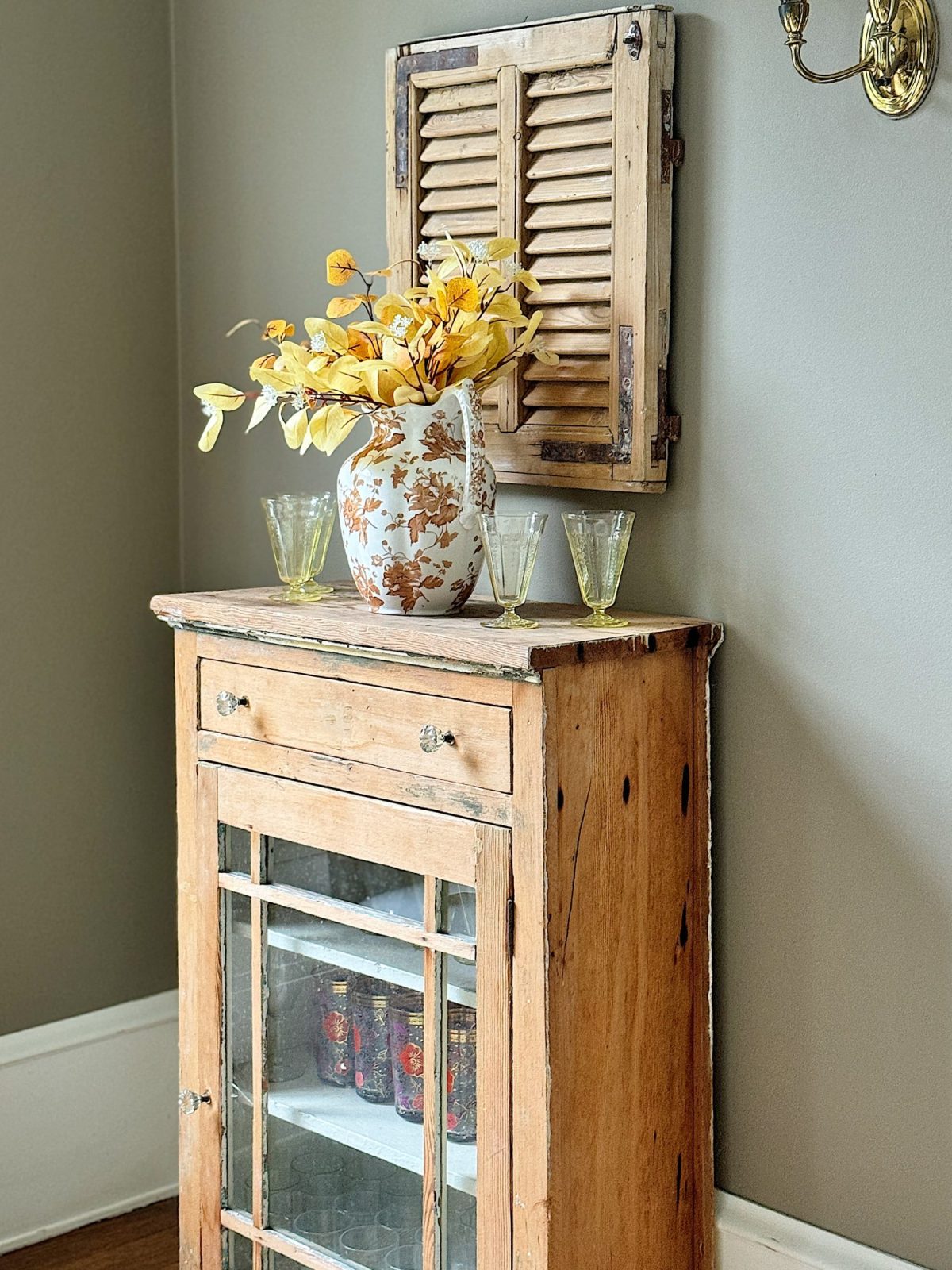 A wooden cabinet with a glass-paneled door holds jars and glasses. A white vase with yellow flowers sits on top, beneath a small decorative shutter on the wall.