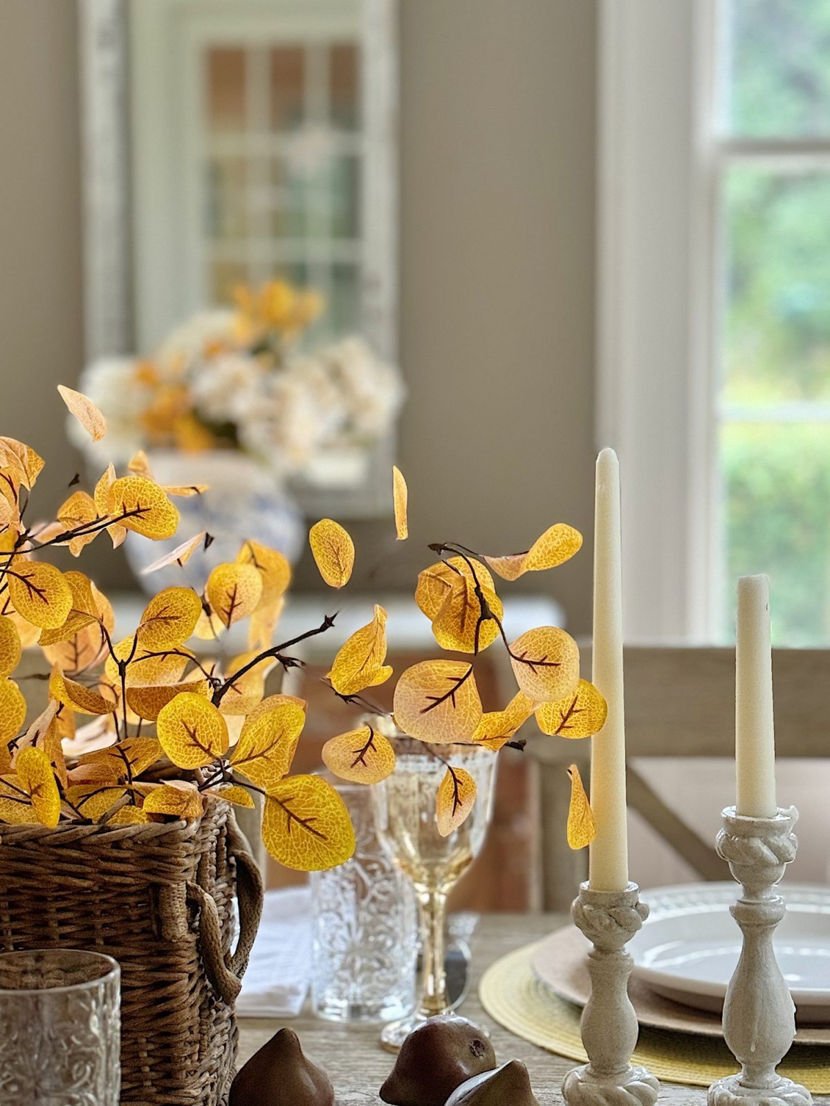 A dining table set with plates, glasses, and candles. A wicker basket with yellow artificial leaves serves as a centerpiece. A mirror and window are visible in the background.