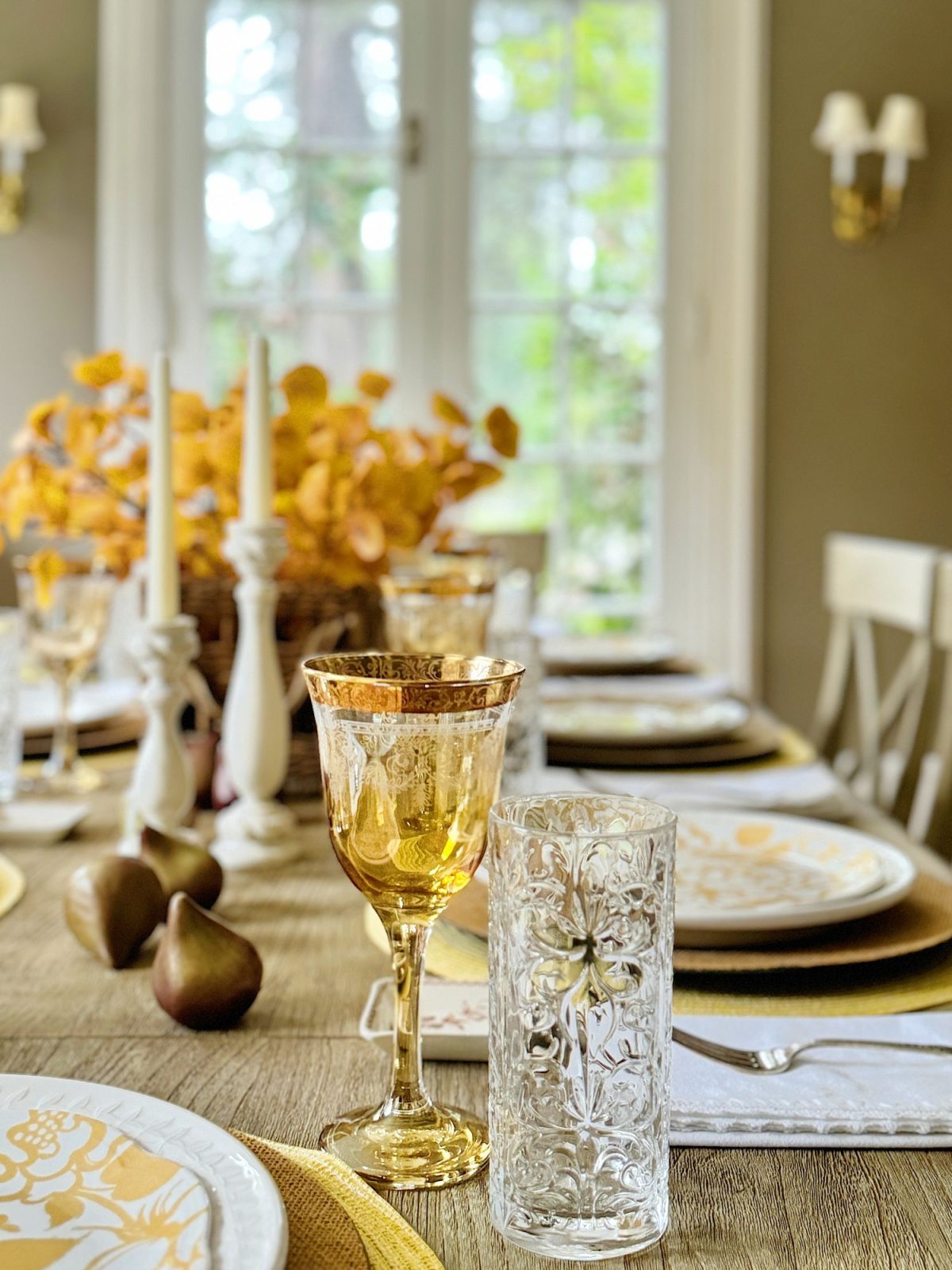 A dining table set with ornate glassware and light yellow dishware, featuring a centerpiece of yellow flowers. Candles and decorative pears add to the elegant arrangement near a window.