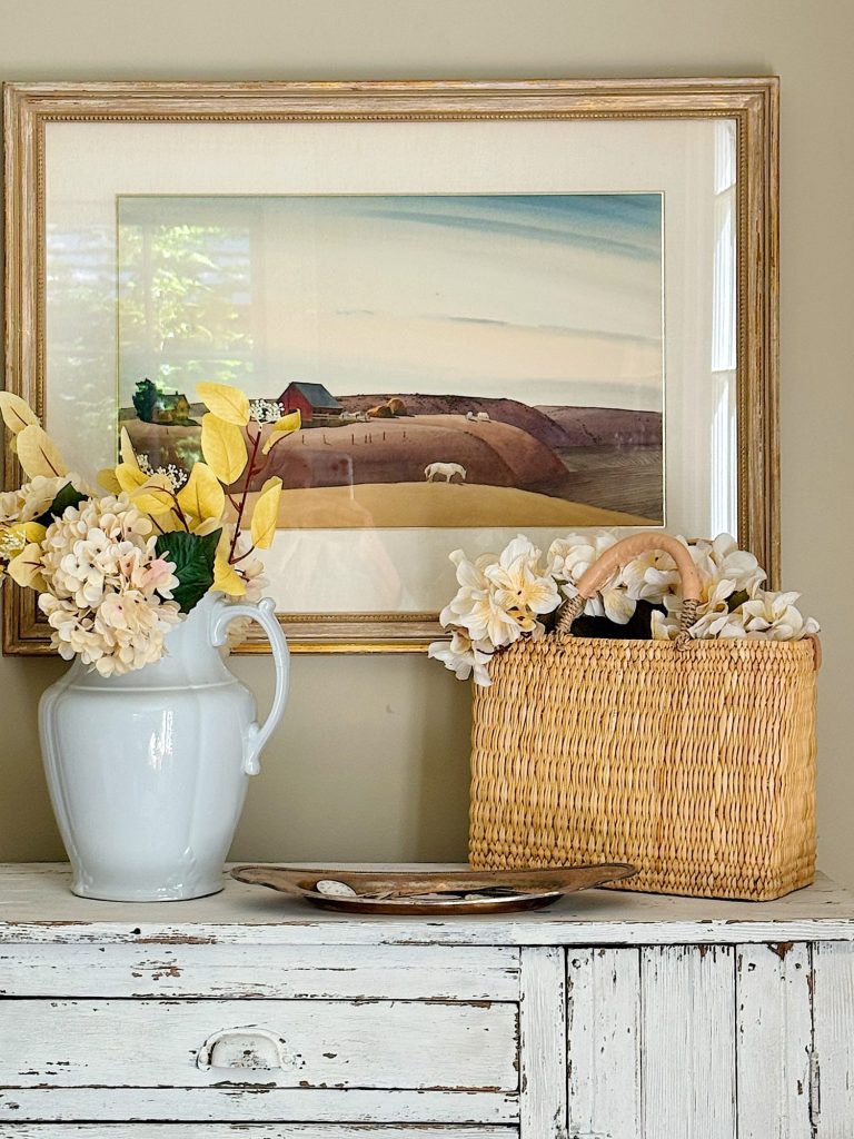 A white ceramic pitcher with yellow and cream flowers and a woven basket with white flowers sit on an antique white dresser. Above them hangs a framed painting of a rural landscape.