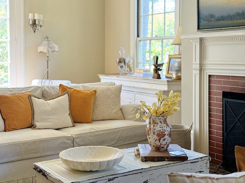 A cozy living room with a white sofa, yellow and beige pillows, a vintage coffee table with a decorative vase and a bowl, a white fireplace, and large windows allowing natural light in.