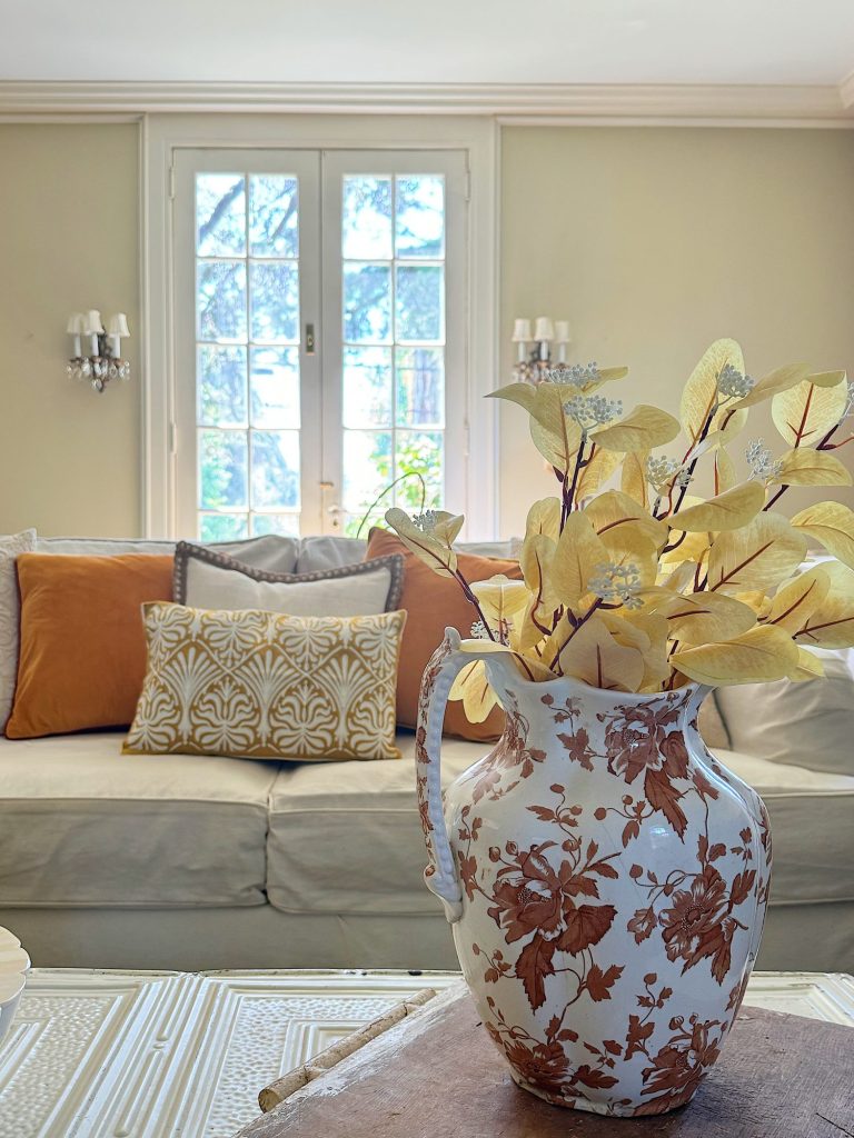 A living room with a white sofa adorned with brown and patterned cushions. A decorative vase with yellow leaves is placed on the coffee table in front of the sofa.