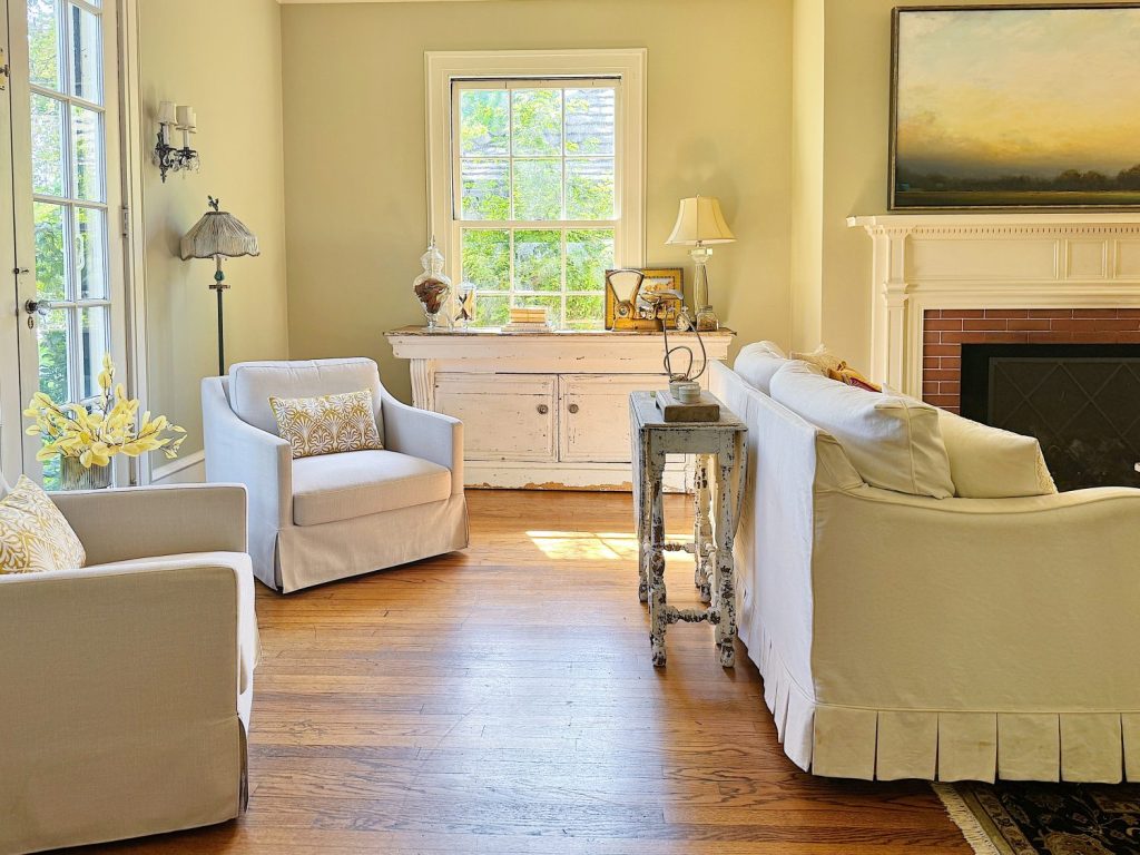 A well-lit living room with light-colored furniture, a white-paneled fireplace, wooden floors, and a large window. A white cabinet with decor items is positioned under the window.