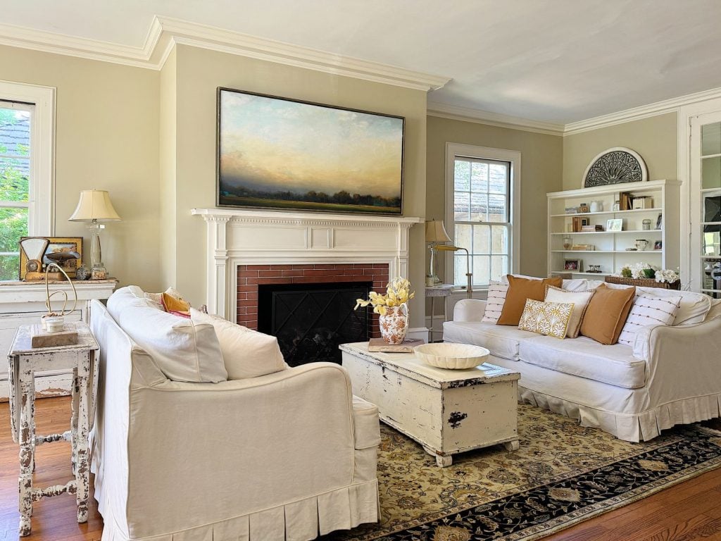 A bright living room with two white sofas, a wooden coffee table, a fireplace with a landscape painting above, and built-in shelves filled with decor items.