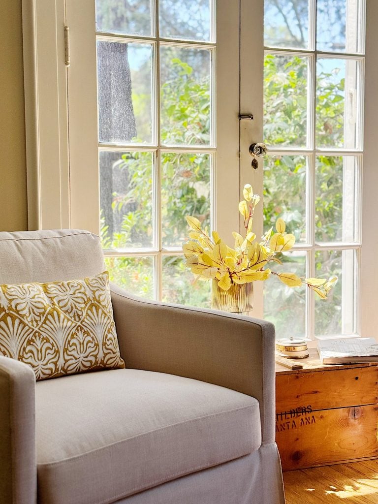 A beige armchair with a patterned pillow is placed next to a wooden chest and a vase of yellow flowers, near a sunlit window with a garden view outside.