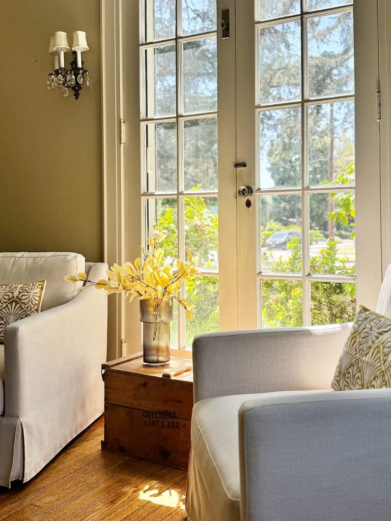 A sunlit living room with white armchairs, patterned cushions, and a glass vase with yellow flowers on a wooden crate by large French doors.