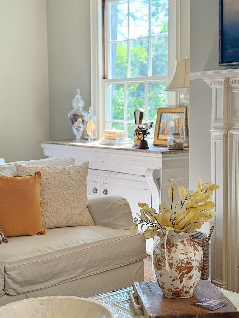 A cozy living room featuring a white couch with pillows, a vase with yellow flowers on a wooden coffee table, a white sideboard with decorative items, and a window providing natural light.