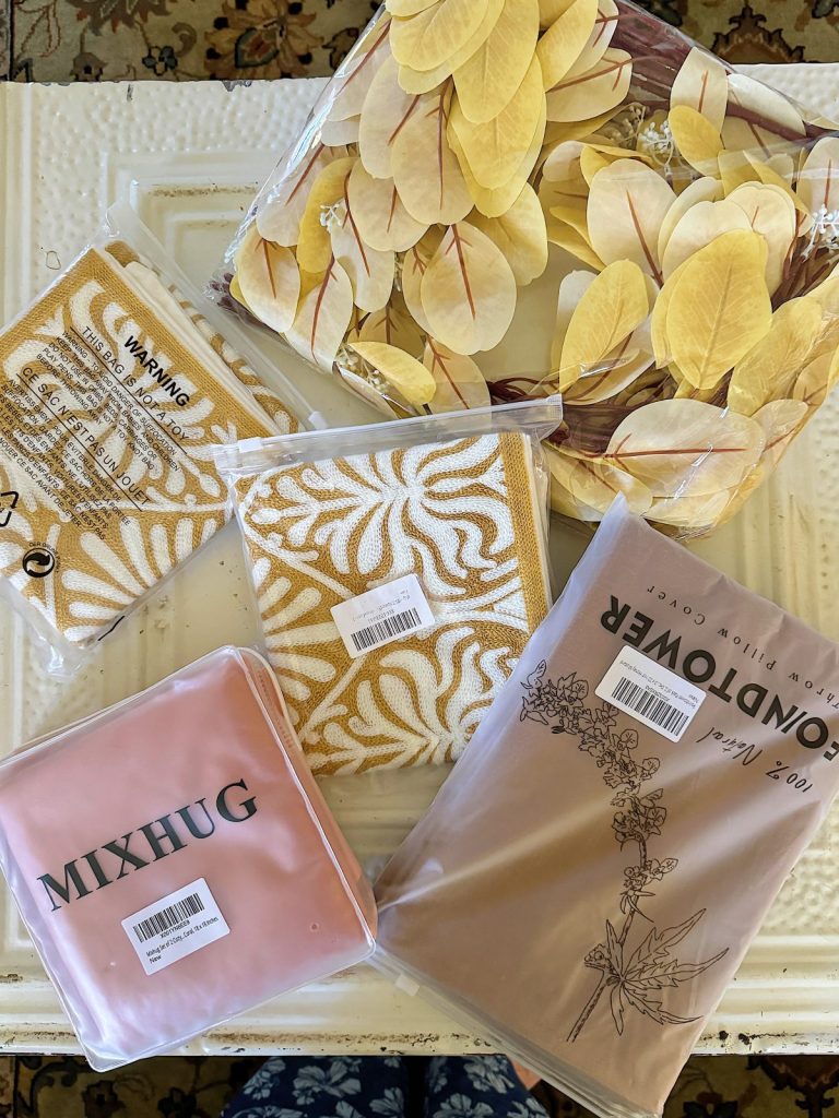 A collection of packaged home decor items, including yellow-patterned textiles, a MIXHUG pink cushion cover, and a BONDTOWER beige pillowcase, displayed on a white table.