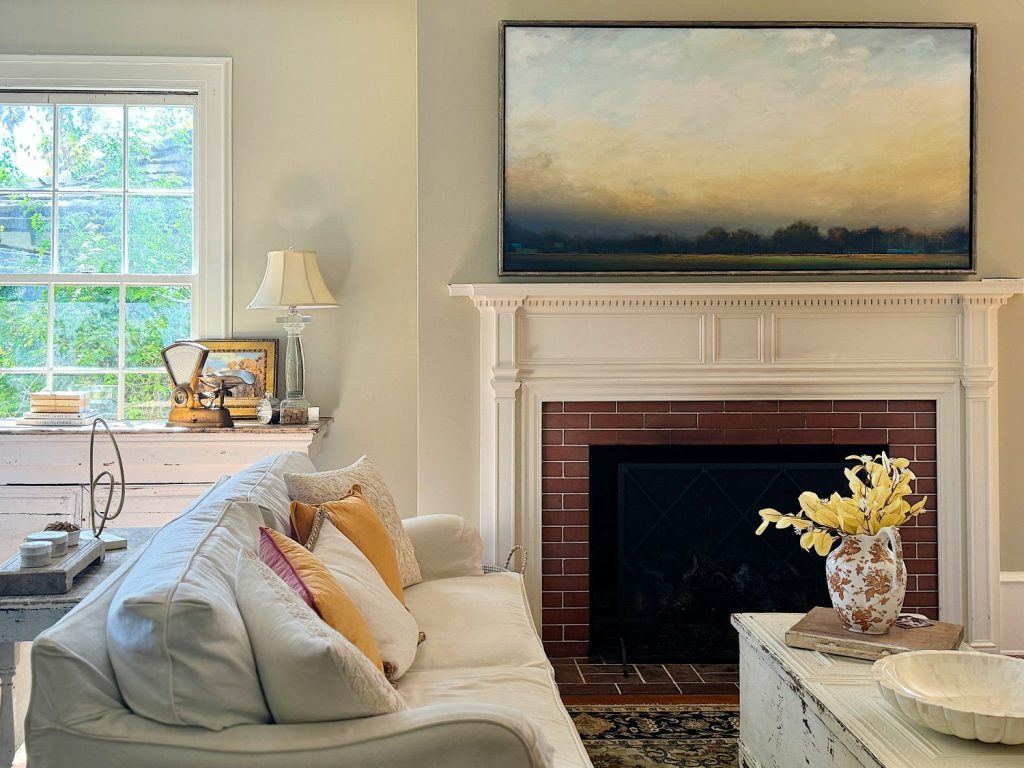 A cozy living room features a white sofa with cushions, a fireplace with a large painting above it, a side table with a lamp, and a window letting in natural light.