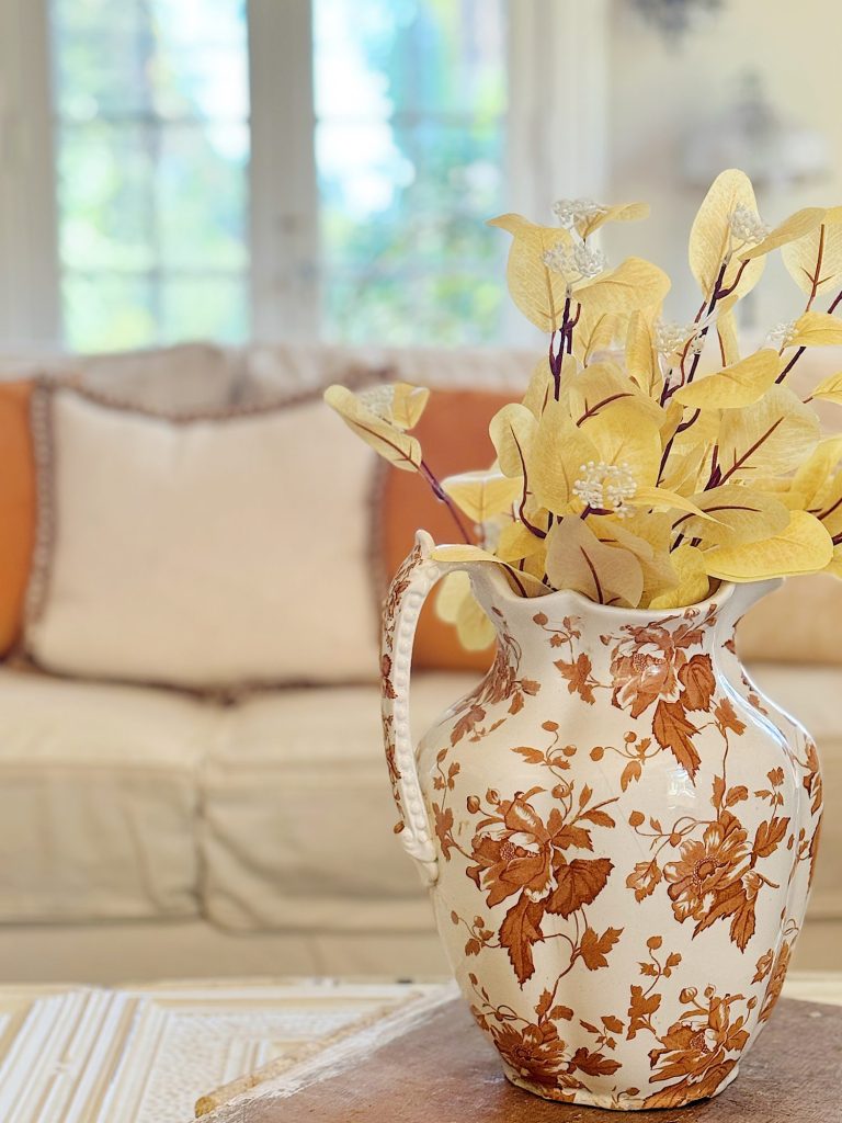 A ceramic pitcher with floral designs holding dried yellow leaves sits on a wooden table in front of a beige sofa with orange and white cushions.