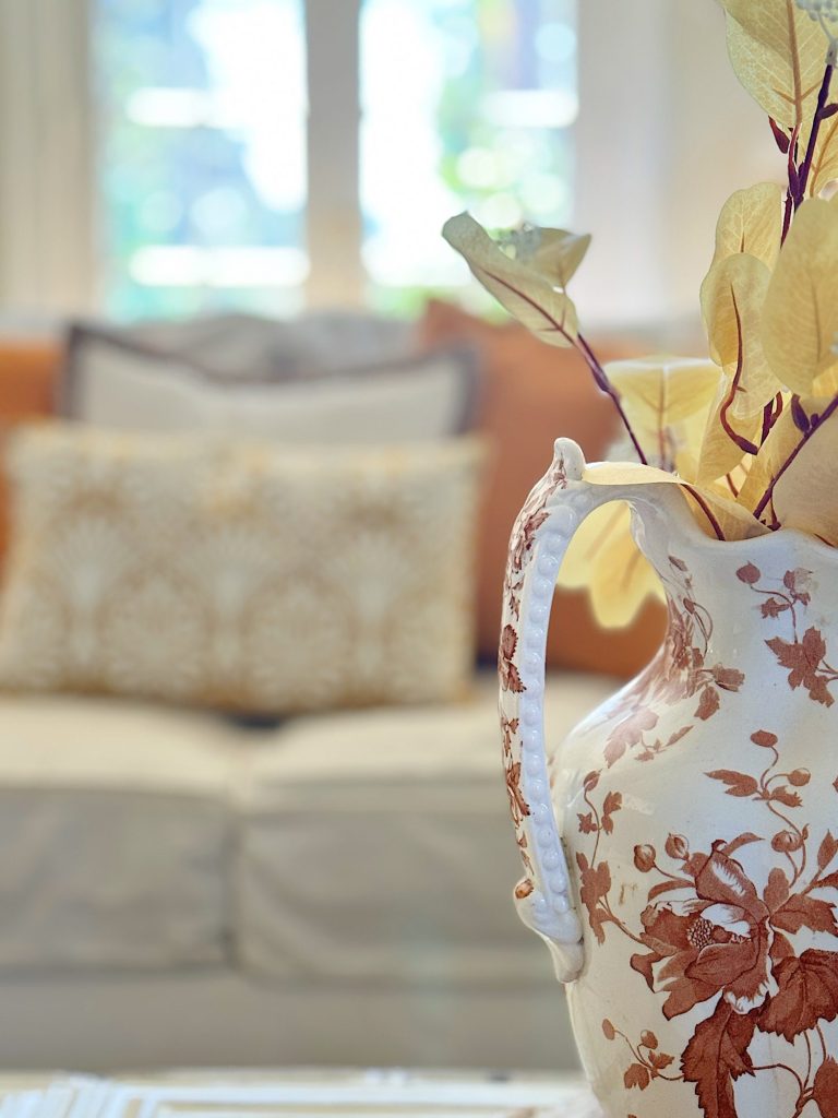 A decorative ceramic pitcher with floral designs, holding dried leaves, is placed in the foreground with a blurred view of a couch and cushions in the background.