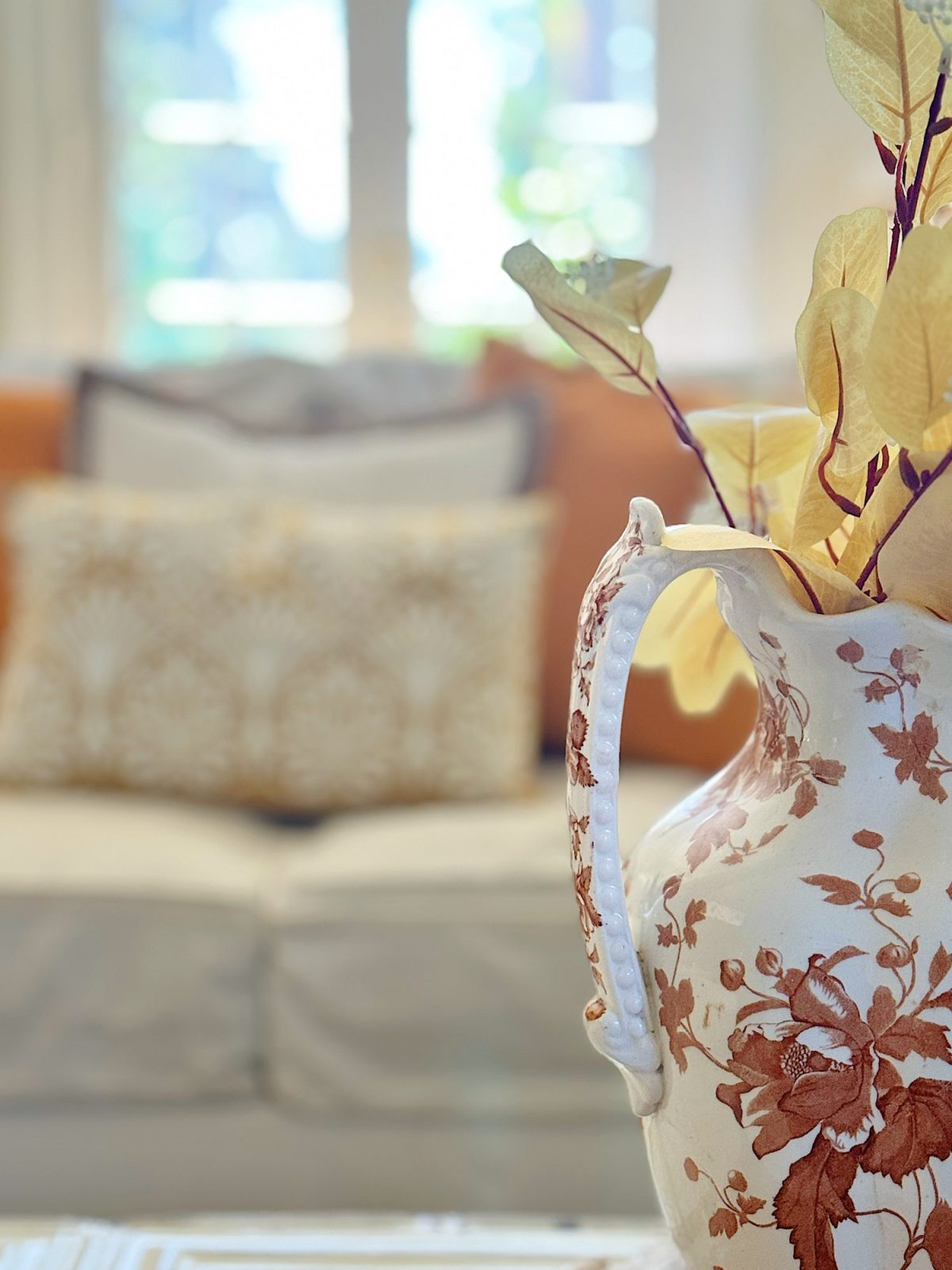 A decorative ceramic pitcher with floral designs, holding dried leaves, is placed in the foreground with a blurred view of a couch and cushions in the background.