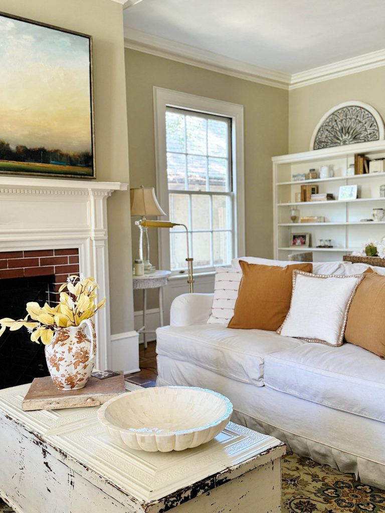 A living room with a white couch, brown and white pillows, a large window, a fireplace with a painting above it, and a distressed coffee table with a decorative bowl and vase of yellow flowers.