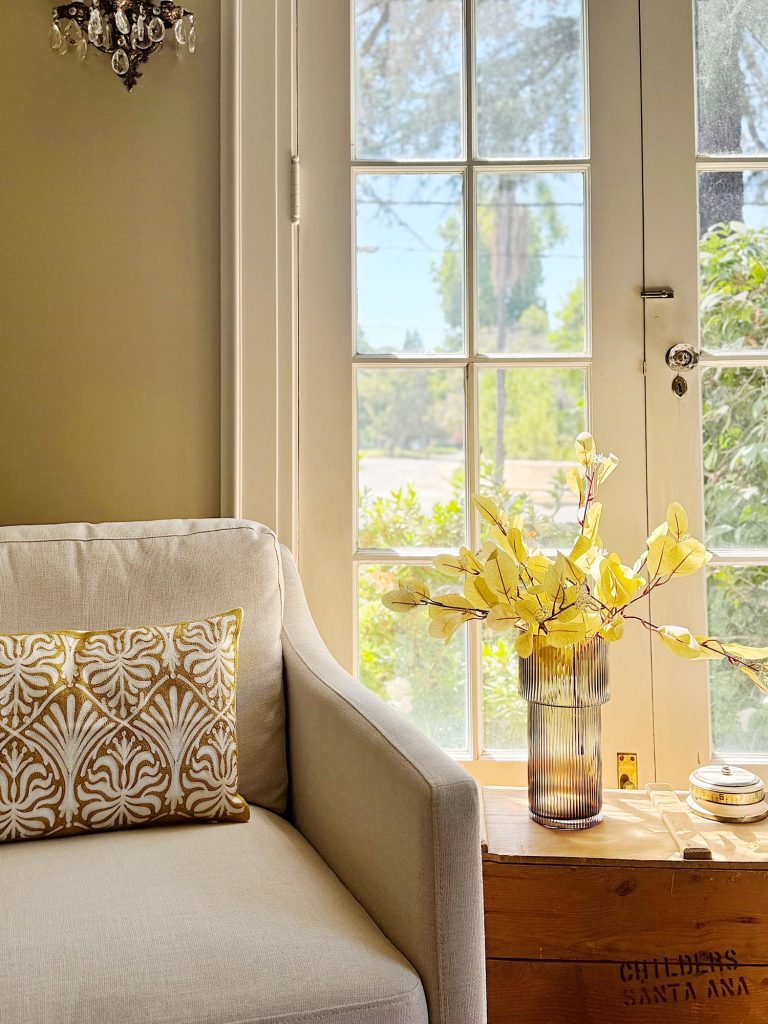 A cozy room with a cream armchair, a decorative pillow, a small wooden crate, a grey vase with yellow leaves, and a glass door leading to an outside view of trees and a bright sky.