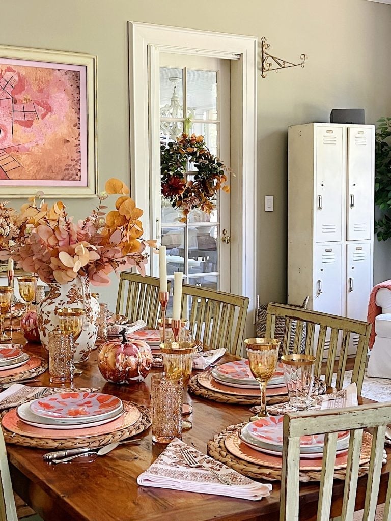 A dining table set with autumn-themed decor, featuring orange and pink plates, amber glassware, and fall foliage centerpieces. A wreath hangs on a glass door in the background, and lockers are placed nearby.