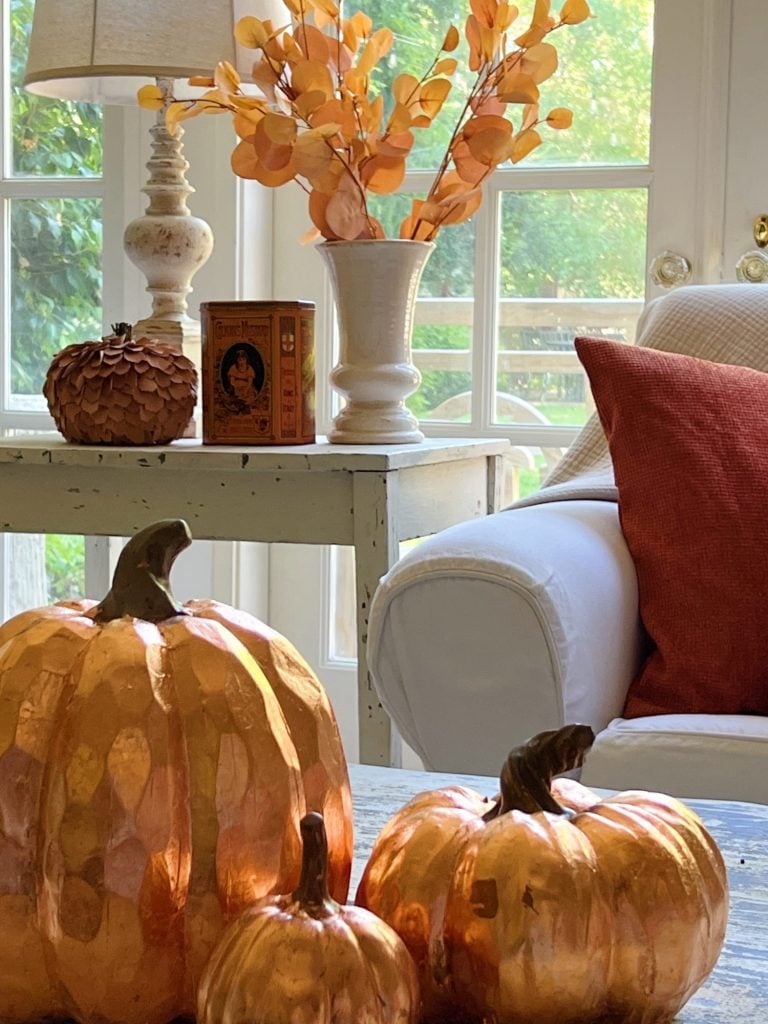 A cozy sitting area with a white armchair, autumn-themed decor including a lamp, vase with orange leaves, pumpkins, and a vintage book, near a window with sunlight streaming in.