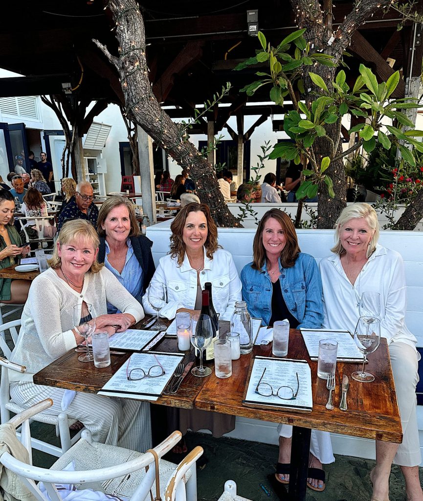 Five women are seated around a wooden table at an outdoor restaurant. They are smiling, and each has a menu in front of them. Drinks are on the table, and trees and other diners are in the background.