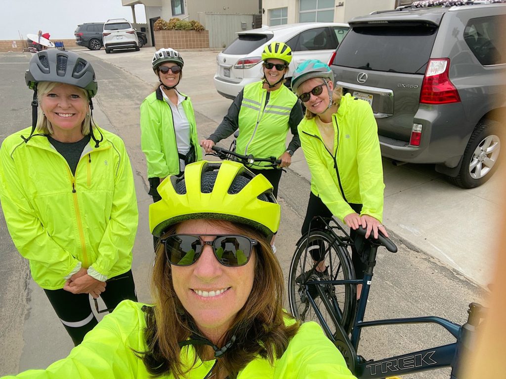 Five cyclists in bright yellow jackets and helmets smile for a group photo on a residential street, with houses and parked cars in the background. One woman holds a bicycle.