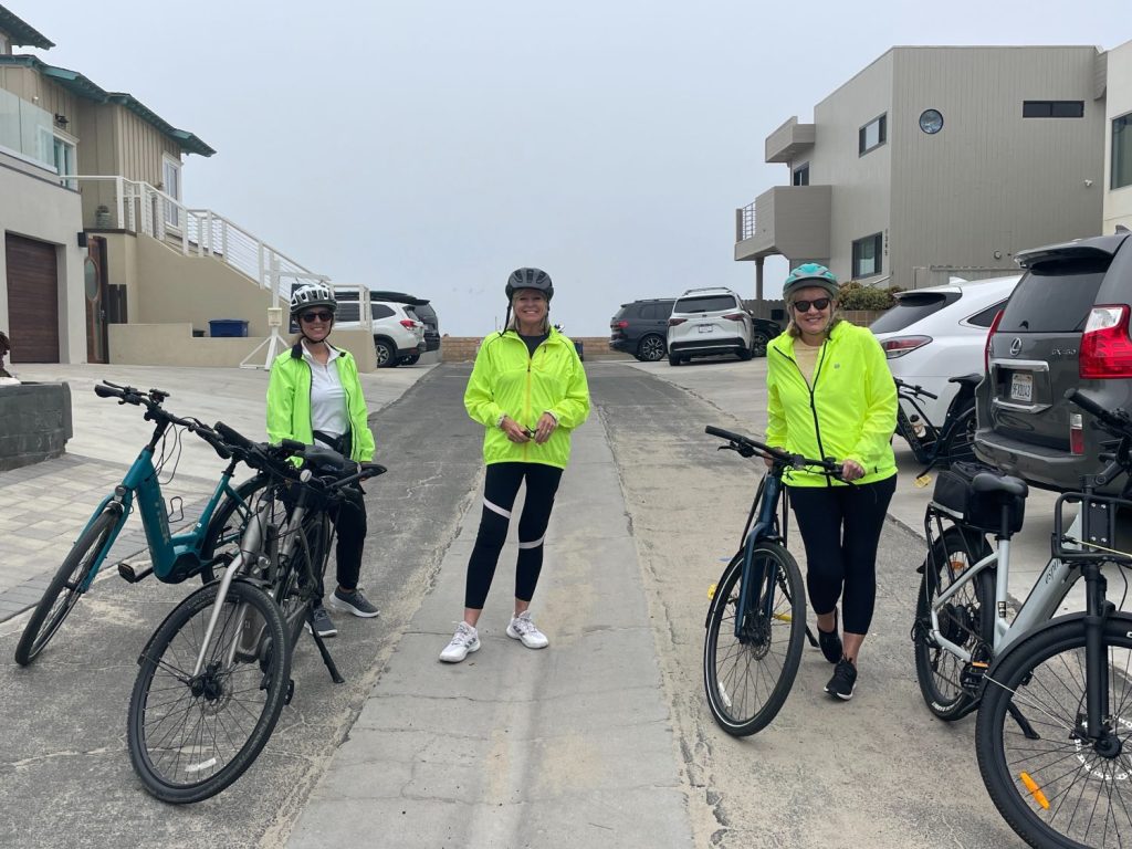 Three people in bright green jackets and helmets stand with their bicycles on a residential street near the beach.