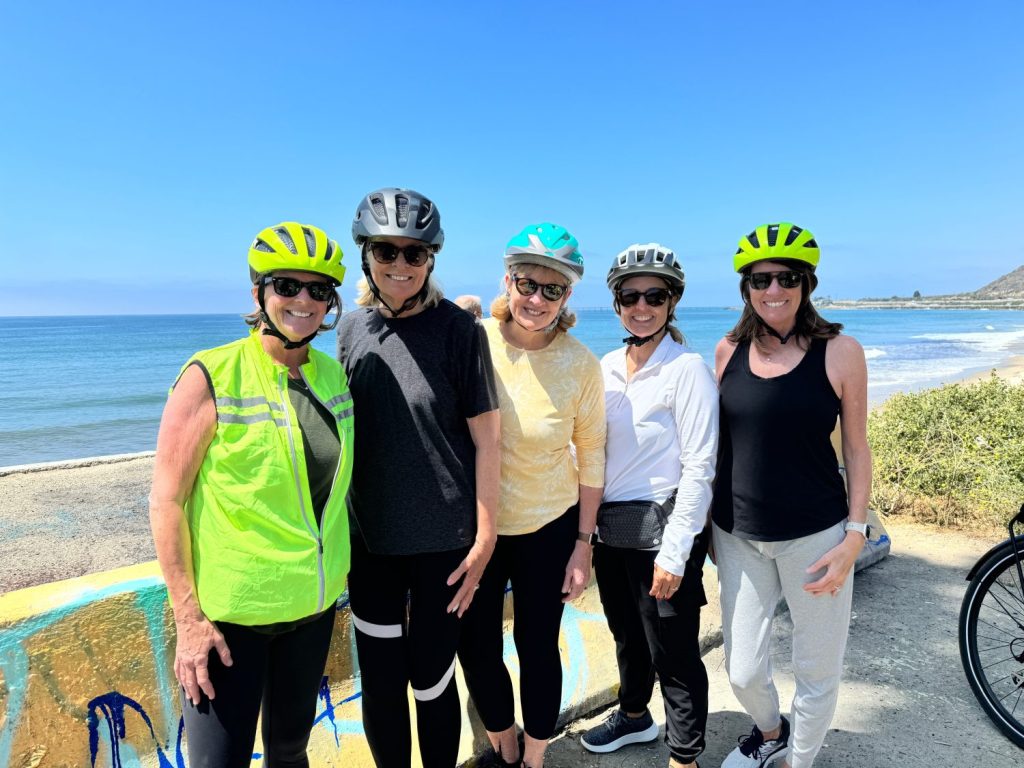 Five women wearing cycling helmets stand together on a coastal path with the ocean in the background.