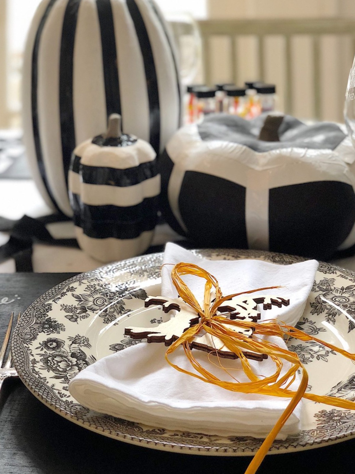 A table setting featuring a decorative plate with a folded white napkin tied with an orange ribbon, surrounded by black and white pumpkins and a fork placed on the left side of the plate.