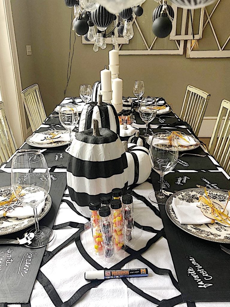 A dining table is set with a black and white Halloween theme, featuring striped pumpkins, candles, wine glasses, and black napkins with messages. Small decorative tubes filled with candy are placed on the table.