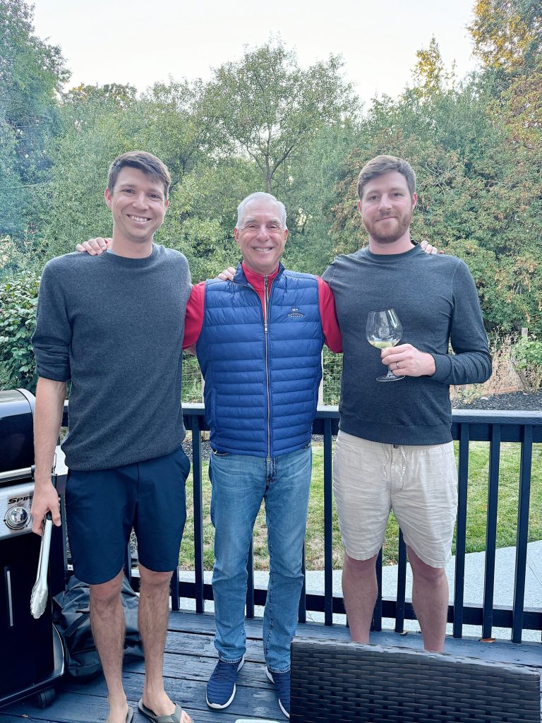 Three men standing on a deck with trees in the background. The central man is wearing a blue vest and red shirt, flanked by two men in gray tops, one holding a glass of wine and a grilling tool.